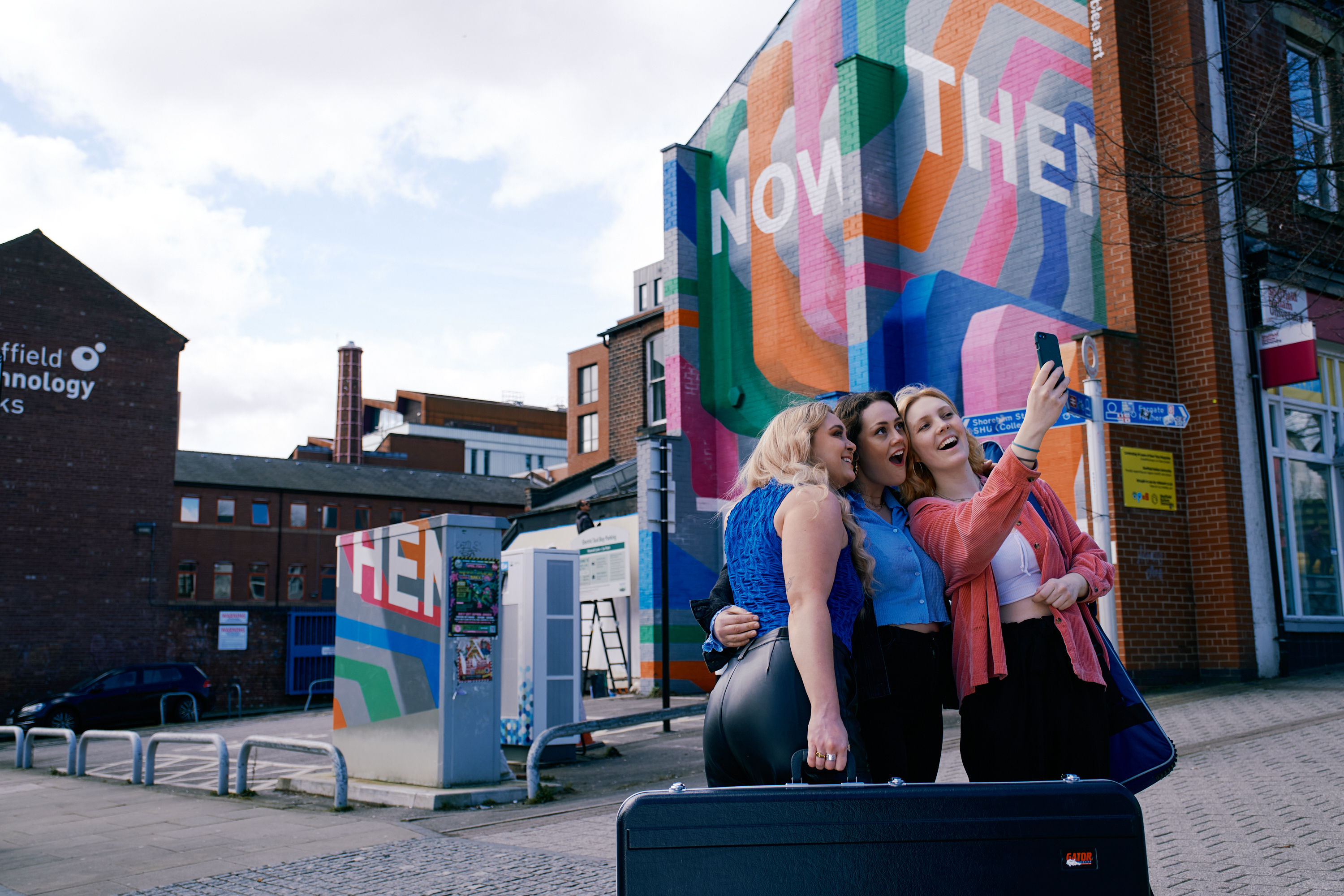 Three women, one of whom is holding a guitar case, pause to take a selfie in front of the Now Then mural on Howard Street in the centre of Sheffield.