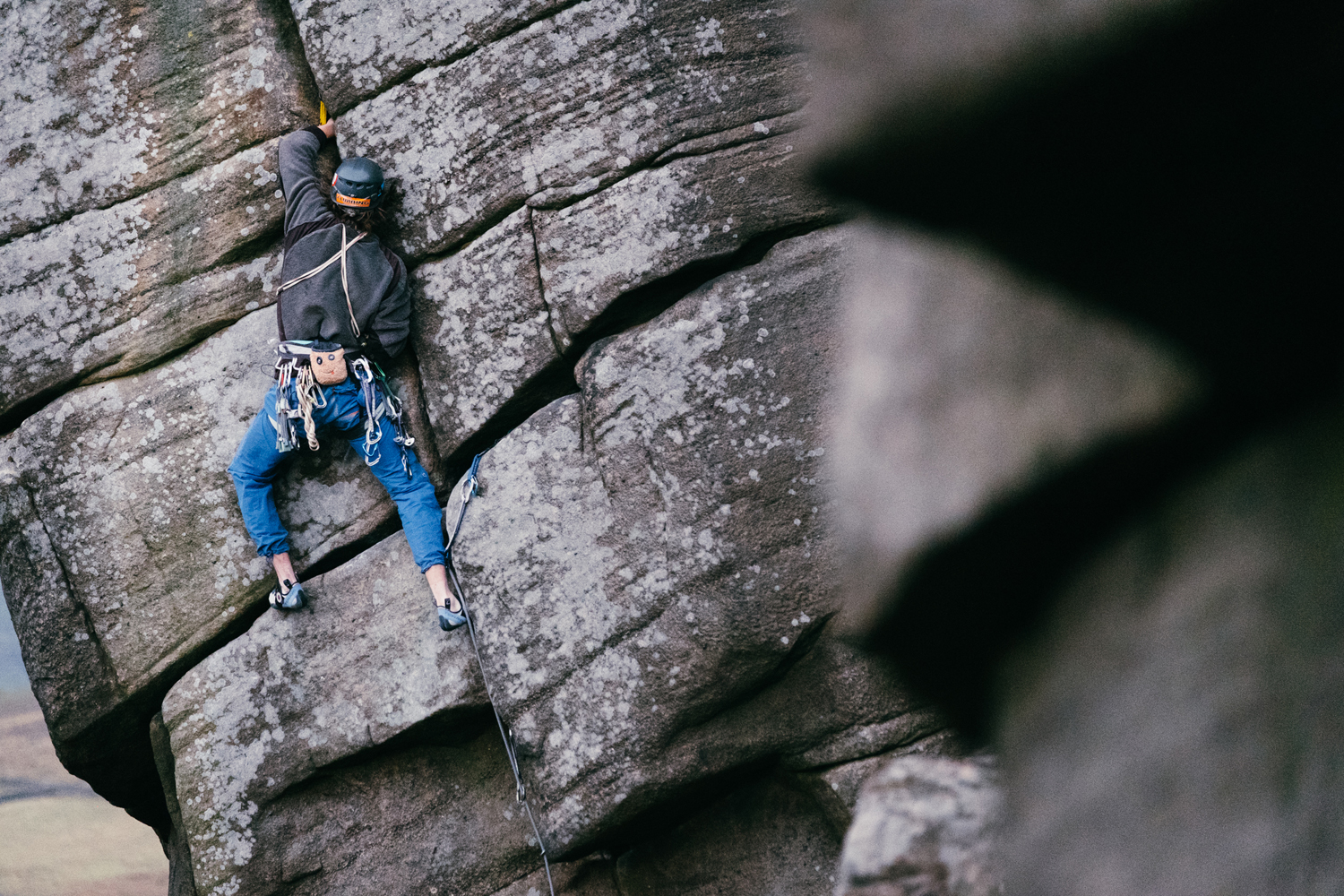 A person wearing a climbing harness and helmet is scaling a large rock face with visible cracks and textured surfaces. The climber is using ropes and safety gear while gripping the rock, and the background shows more rugged stone formations.