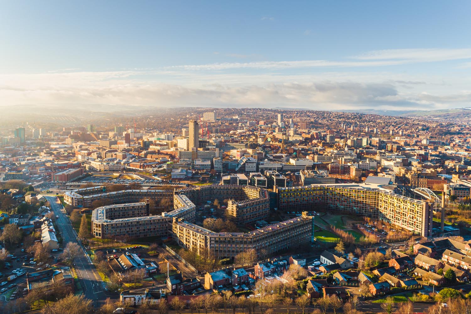 Aerial view of Sheffield cityscape under clear skies, showing a mix of modern and historic buildings. Prominent in the foreground is the curved Park Hill housing complex with its distinctive architecture, surrounded by roads and greenery. The city centre stretches into the distance with tall buildings, including the central tower, and rolling hills visible on the horizon.