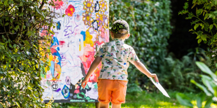 A child is walking along a sunlit woodland path surrounded by dense green foliage, holding a piece of paper in one hand. Ahead on the path is a large, brightly coloured abstract artwork propped up outdoors, featuring splashes of pink, yellow, blue, and various shapes. Sunlight filters through the trees, creating dappled light on the ground and highlighting the vibrant scene. James Mulkeen for WWPT