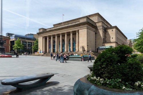 Large neoclassical building with tall columns and steps at the entrance, located in a spacious paved square. Several people are walking or standing in front of the building, and greenery with flowers is visible in the foreground. Modern buildings and trees surround the square under a clear blue sky.