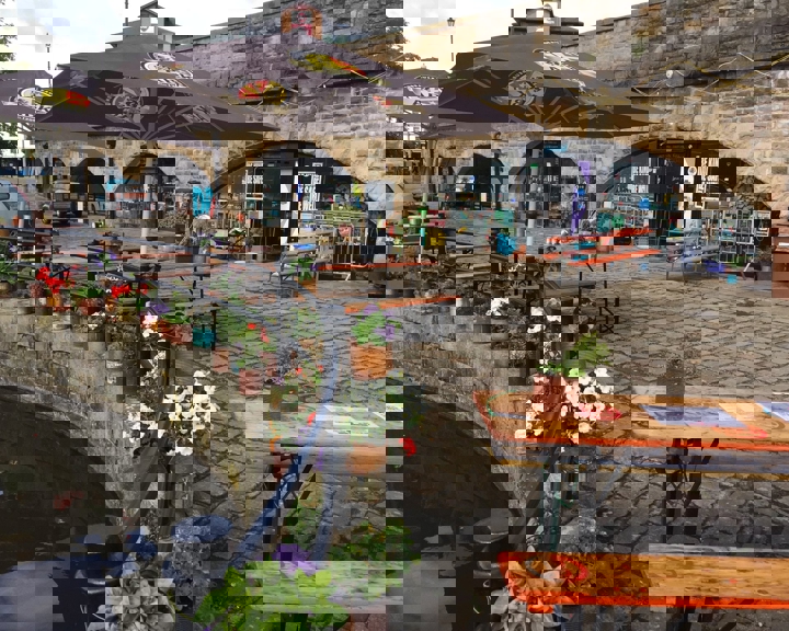 Outdoor seating area in front of a stone building with large archways. The space features wooden picnic-style tables with orange tops & green metal legs, some under large branded umbrellas. Colourful potted flowers line the edge of a canal in the foreground, adding vibrant reds, whites, and purples. Behind the seating area, the building has signage and posters visible through the arches, suggesting a café or bar setting. The ground is paved with cobblestones, and the overall scene has a relaxed atmosphere.