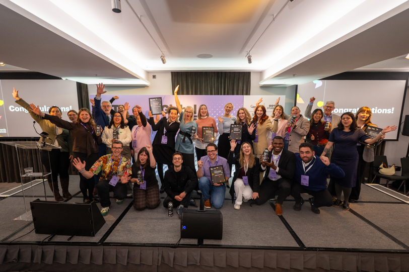 A large group of people gathered on a stage in a conference-style room, posing together with raised arms and award plaques. The setting includes wide projection screens displaying celebratory messages and bright lighting that highlights the event’s festive atmosphere.