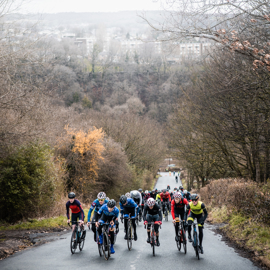A group of cyclists riding uphill on the road, with part of the city in shot in the distance.