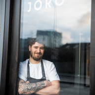 A person wearing a white short-sleeved chef’s jacket and a dark apron stands with arms crossed inside a glass doorway. The glass reflects an urban street scene with buildings and a cloudy sky. Visible text on the glass reads “0.3 JÖRO” in white letters near the top. The individual has a tattoo sleeve on one arm, and the setting suggests the entrance to a modern restaurant.