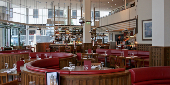 The interior at Piccolino. Bright, modern restaurant interior with large windows, high ceilings, and hanging pendant lights. The space features curved wooden booths with red leather seating and neatly set tables with glassware and menus. A long bar at the back is stocked with bottles, and the overall design combines warm wood tones with contemporary elements.