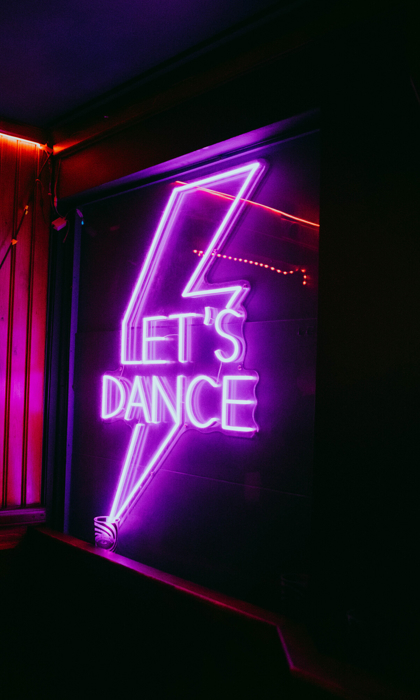 Pink and purple neon sign reading “Let’s Dance” inside a dimly lit bar or club space.