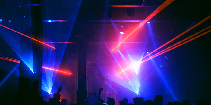 A crowded nightclub or music venue with vibrant laser lights in blue and red cutting across the dark space. Silhouettes of people are visible in the foreground, some with raised arms, creating a lively atmosphere. The beams of light converge toward the center stage area, highlighting the industrial-style ceiling and pillars. The overall scene conveys an energetic party or concert setting with dramatic lighting effects.