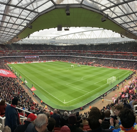 Panoramic view of a packed football stadium with fans filling the stands. The green pitch is set for a match, with goalposts visible at both ends. Large red banners and flags are displayed by supporters, and the stadium roof arches overhead with a modern design.