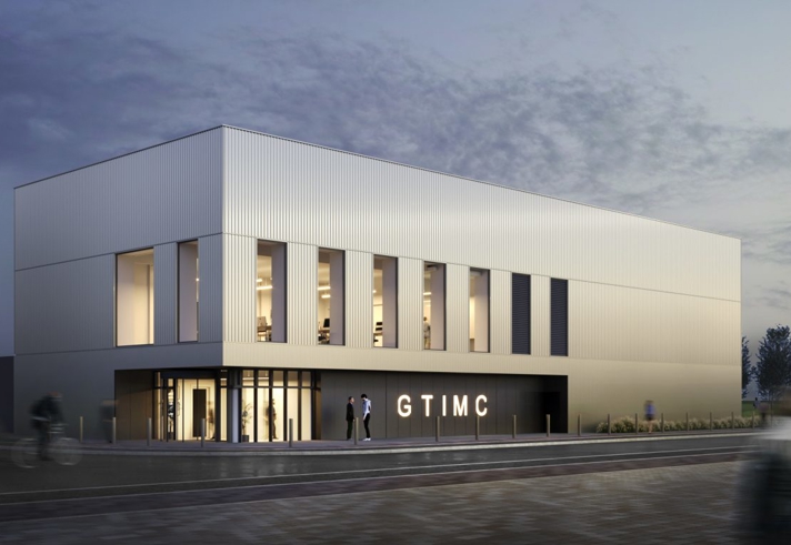 The exterior of the Gene Therapy Centre building. Modern two-story building with a sleek, minimalist design featuring vertical metal panels and large rectangular windows. The structure is illuminated from inside, creating a warm glow against the twilight sky. The entrance has glass doors and signage reading “GTIMC.” A few people are visible near the entrance, and blurred motion suggests activity in the foreground.