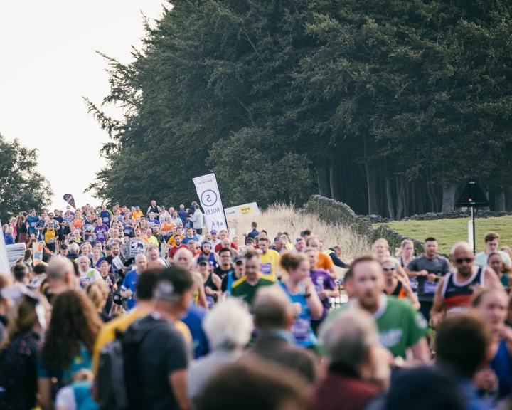 A large crowd is watching and cheering on runners running on a road. In the background is a large wood.