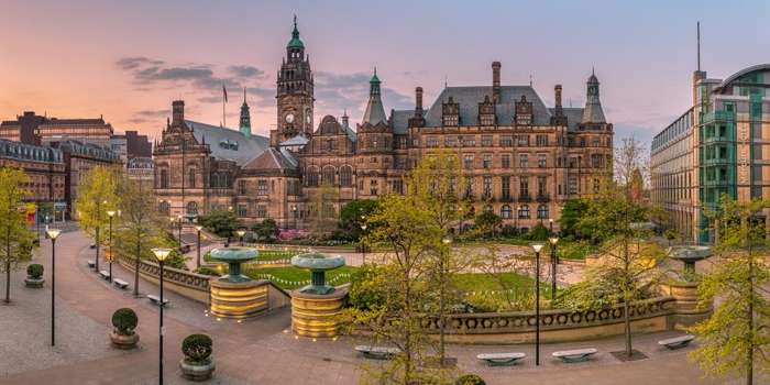 The Peace Gardens in Sheffield city centre; Panoramic view of a historic city square at sunset, featuring a large ornate building with towers and detailed architecture, likely a town hall. The square includes landscaped areas with trees, benches, and tiered fountains. Modern buildings border the scene, and the sky glows with warm orange and pink tones.