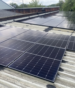 Rows of black solar panels installed on a corrugated metal rooftop. The panels are arranged in neat grids, reflecting light under a cloudy sky. In the background, there are additional rooftops, trees, and a glimpse of the horizon.