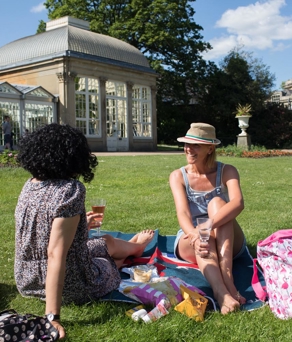 Two women are sat on the grass, on a bright, sunny day, at The Sheffield Botanical Gardens having a picnic and chatting. 