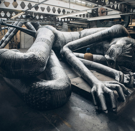 A large, intricate sculpture of a reclining humanoid figure with elongated limbs and detailed patterns covering its surface lies on the floor of an industrial-style warehouse. The artwork features exaggerated hands and feet, a textured body, and a stylized head with decorative markings. Overhead, metal beams and geometric cutouts line the ceiling, while machinery and pipes are visible in the background.