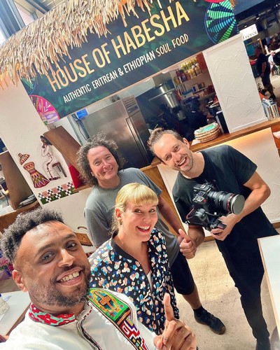 Group of people standing inside a food stall named ‘House of Habesha,’ which has a sign reading ‘Authentic Eritrean & Ethiopian Soul Food.’ The stall features cultural artwork on the walls, and one person is holding a professional camera.