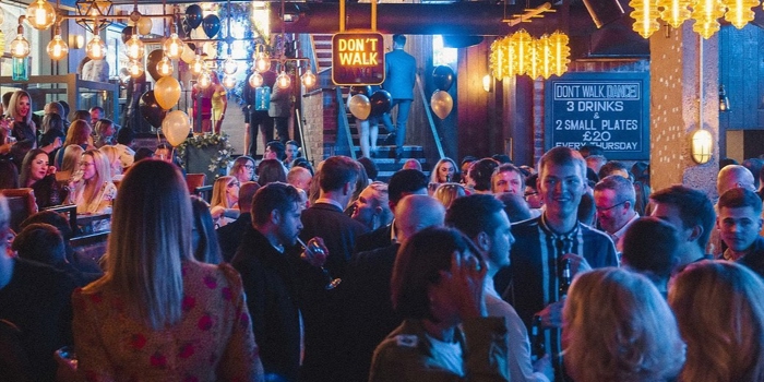 Manahatta filled with people. Crowded bar scene with a large disco ball hanging from the ceiling, surrounded by warm pendant lights and gold balloons. People are gathered closely, chatting and holding drinks. A neon-style sign reading “DON’T WALK” hangs above the crowd, and a chalkboard menu on the right advertises drink and food specials. The atmosphere is lively with dim, colourful lighting and industrial-style decor.