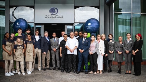 Group of people standing in front of the entrance to a DoubleTree by Hilton hotel, posing for a photo. The group includes individuals wearing business attire and others in casual uniforms, with two large branded balloons on either side of the doorway.