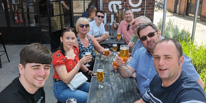A group of people sat round a table, outside a restaurant, drinking beers.
