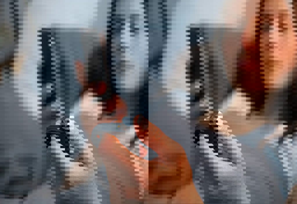 Close-up of a person lying in bed holding a smartphone in one hand and a small black handheld device in the other. The person is covered with a gray blanket, and the background shows pillows and bedding.