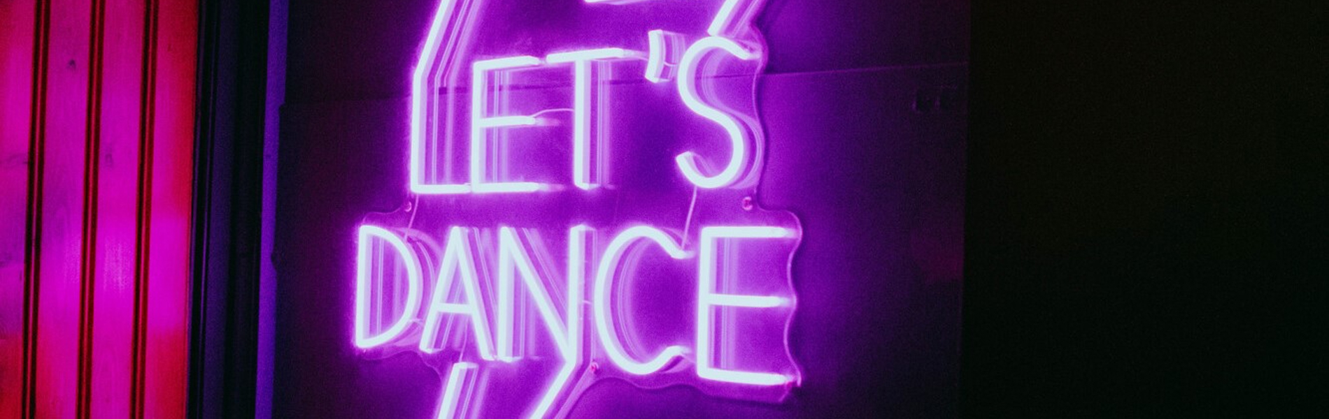 Pink and purple neon sign reading “Let’s Dance” inside a dimly lit bar or club space.