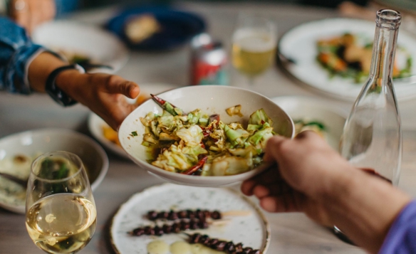 A close-up of a dining table with several dishes and drinks. In the foreground, two hands are passing a white bowl filled with a vegetable dish that includes chopped greens and red chili peppers. Surrounding the bowl are plates with various foods, a glass of white wine, a clear glass bottle, and a can of soda. The table setting suggests a shared meal in a casual, social atmosphere.