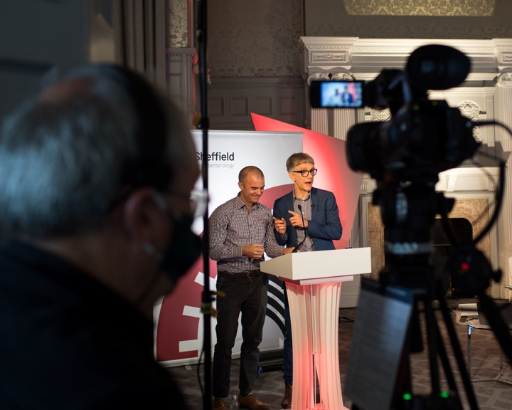 Two presenters standing behind a white podium on a stage, speaking into microphones during an event. A professional video camera on a tripod is positioned in the foreground, filming the scene. The backdrop includes event branding with the word ‘Sheffield’ partially visible, and the setting features ornate wall panelling and decorative architecture.