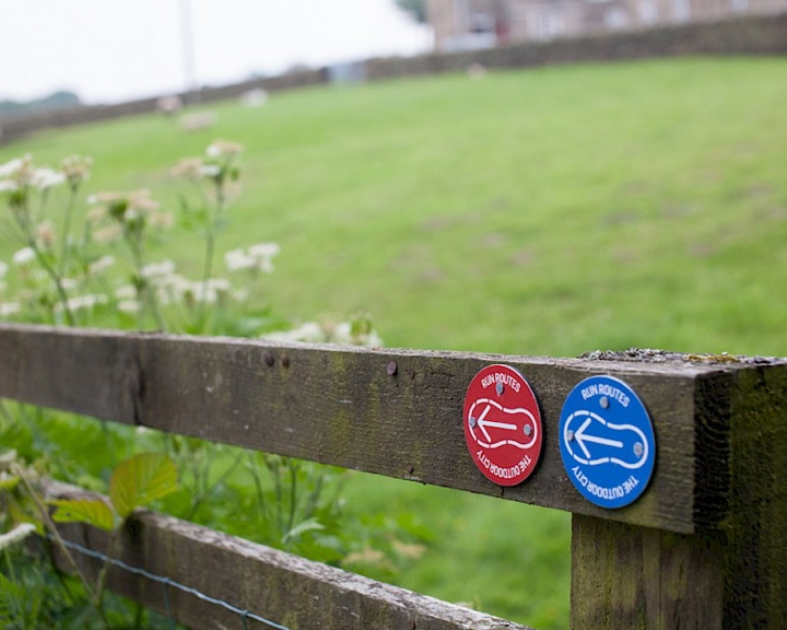 A wooden fence with two circular trail markers attached to it, one red and one blue, each showing a white arrow symbol. Behind the fence is a grassy field with wildflowers in the foreground and a stone wall in the distance.
