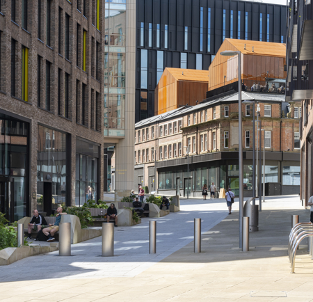 A pedestrianised shopping street in a city centre. People are strolling up and down, while others are sitting on benches.