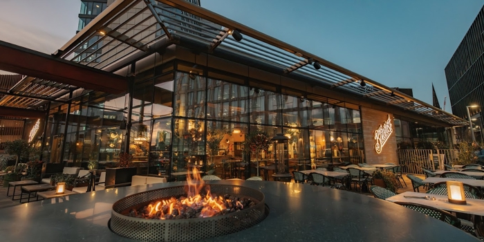 An evening view of a glass-walled restaurant surrounded by outdoor seating, with a circular fire pit lit in the foreground and soft lighting throughout the terrace.
