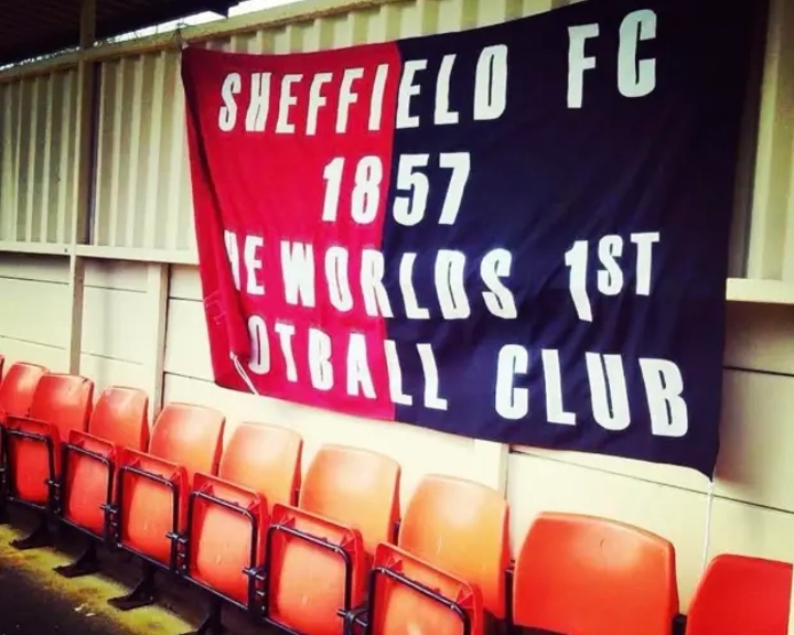 A banner hanging on a corrugated wall behind a row of red stadium seats. The banner is split into red and black halves and reads in bold white letters: ‘Sheffield FC 1857 The World’s 1st Football Club.’