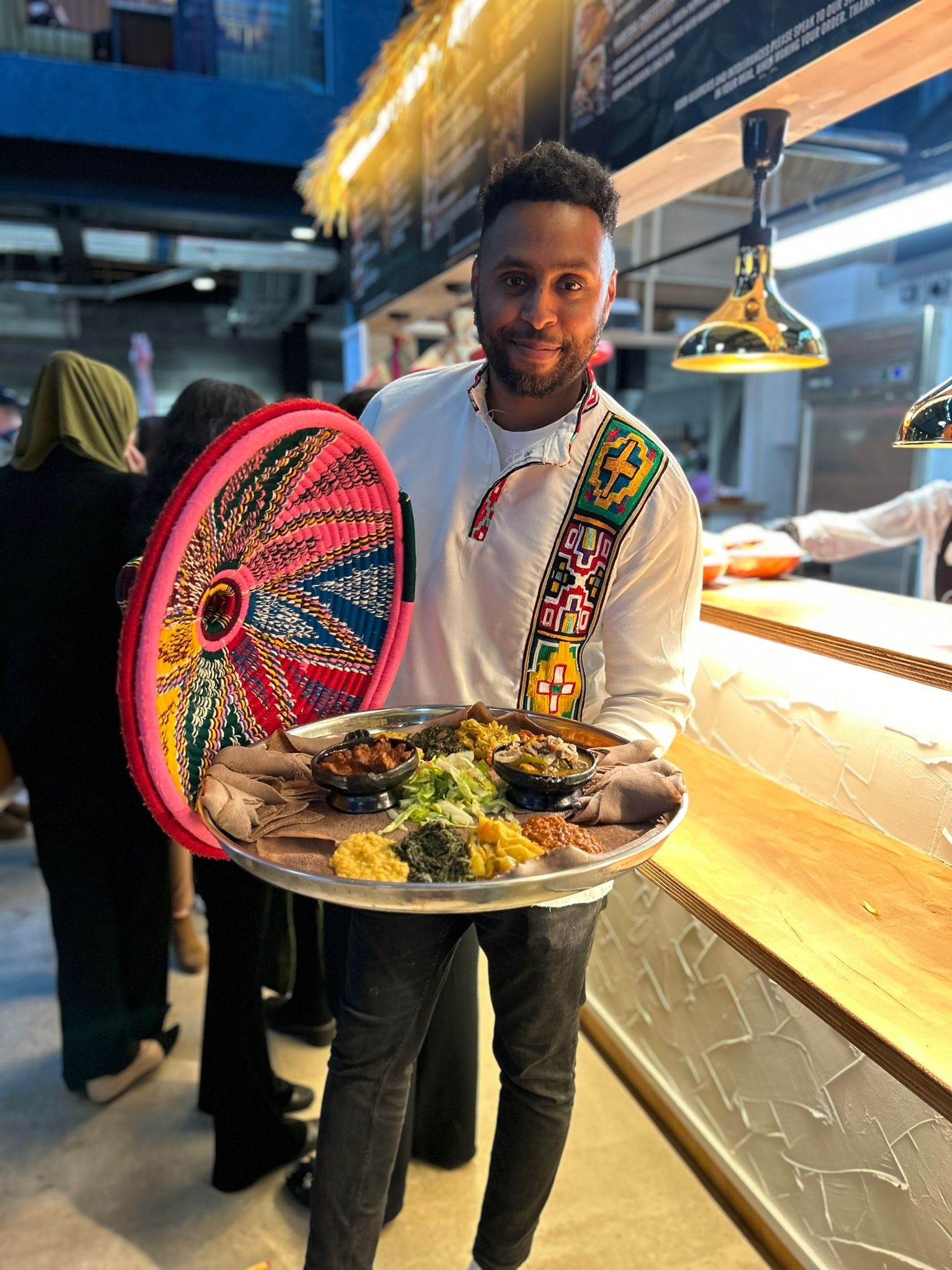 Person wearing a white traditional-style shirt with colorful embroidered patterns, holding a large round woven basket lid and a metal tray filled with Ethiopian dishes. The tray includes injera topped with various stews and vegetables. The setting appears to be an indoor food market or restaurant counter with warm lighting and people in the background.