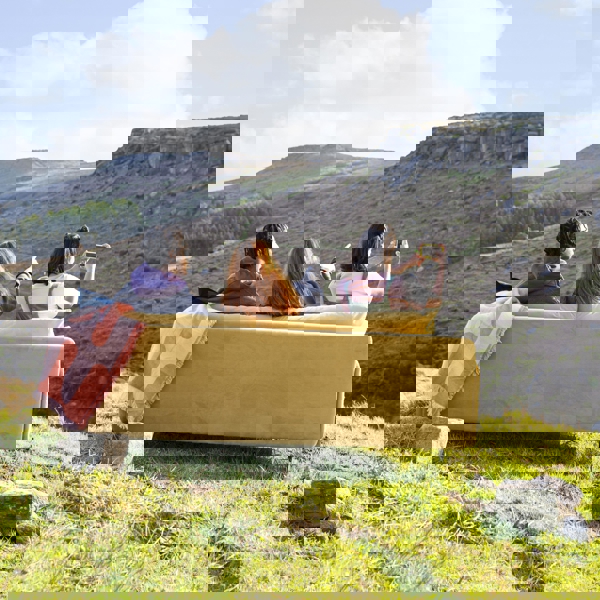Three people sitting on a yellow couch placed outdoors on a grassy hillside, overlooking a rugged landscape with rocky terrain and distant hills under a bright blue sky with scattered clouds. One person is pointing toward the horizon while another holds up a phone, possibly taking a photo.