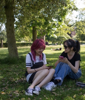 Two people sitting on the grass in a park under the shade of trees. They are surrounded by green foliage and scattered fallen leaves, with one person holding a smartphone. In the background, other people are relaxing on the grass, and the scene is bright and sunny with open green space and trees.