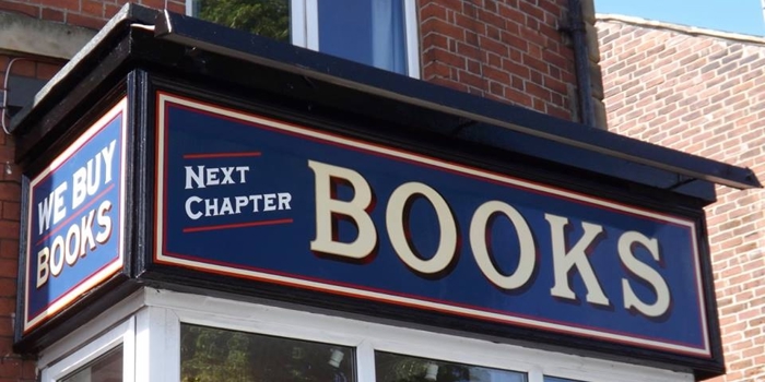 Close-up view of the Next Chapter Books shop sign, featuring bold cream lettering on a dark blue background with red and white borders. The phrases ‘WE BUY BOOKS’ and ‘NEXT CHAPTER BOOKS’ are displayed on the corner fascia of a red‑brick building.