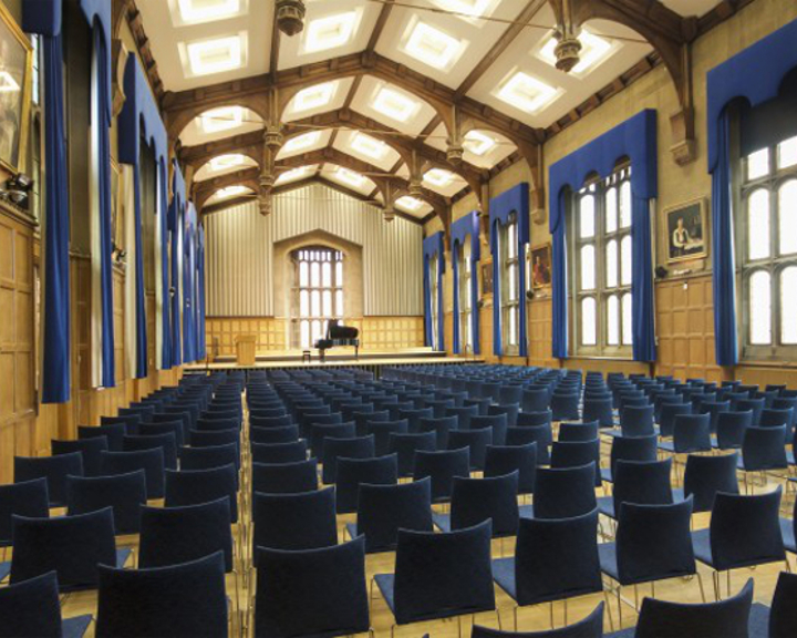 Interior of a grand hall with high wooden-beamed ceiling and large arched windows draped in blue curtains. Rows of blue chairs face a raised stage with a black grand piano, and the walls feature wood panelling and framed portraits.