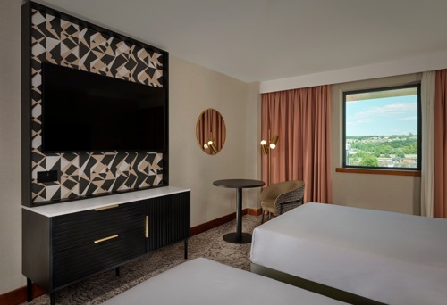 Modern hotel room with two beds, a large flat-screen TV mounted on a patterned panel above a black cabinet, a round mirror on the wall, a small round table with a chair, and a window with pink curtains showing a scenic view outside.