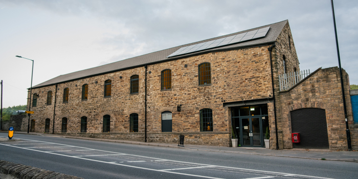 Exterior view of a long stone mill building with arched windows, a pitched roof and a glass‑fronted entrance facing the roadside.