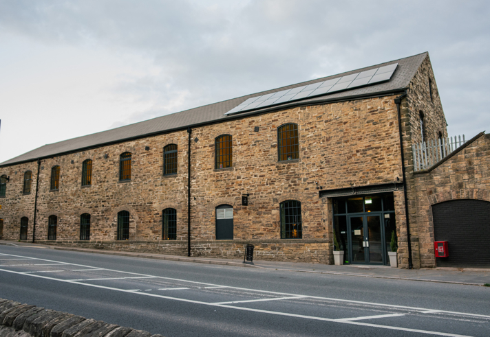 Exterior view of a long stone mill building with arched windows, a pitched roof and a glass‑fronted entrance facing the roadside.