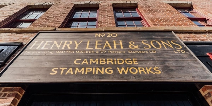 Close-up view of a large wooden sign mounted on a red-brick building. The sign reads: ‘No 20 Henry Leah & Sons Ltd, incorporating Walter Walker & Co Piercers Stampers Ltd, Cambridge Stamping Works.’ The building has tall windows and a clear blue sky above.