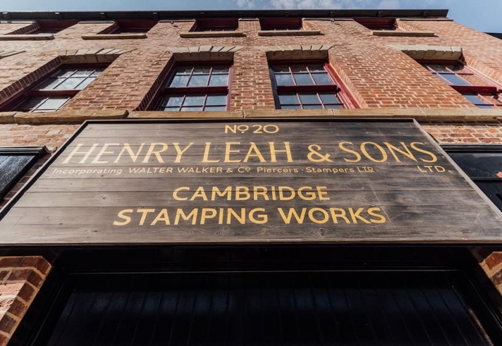 Close-up view of a large wooden sign mounted on a red-brick building. The sign reads: ‘No 20 Henry Leah & Sons Ltd, incorporating Walter Walker & Co Piercers Stampers Ltd, Cambridge Stamping Works.’ The building has tall windows and a clear blue sky above.