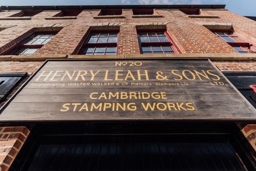 Close-up view of a large wooden sign mounted on a red-brick building. The sign reads: ‘No 20 Henry Leah & Sons Ltd, incorporating Walter Walker & Co Piercers Stampers Ltd, Cambridge Stamping Works.’ The building has tall windows and a clear blue sky above.