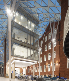 Heartspace - Modern atrium with a glass roof and steel beams connecting to a historic red-brick building. The space features multiple floors with large glass windows, bright lighting, and a seating area with tables and chairs on the ground level
