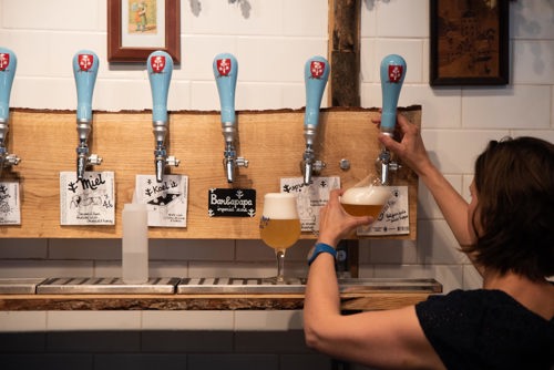 Person pouring a draft beer from a tap in a bar with six blue-handled beer taps mounted on a wooden panel. Two glasses of beer are on the counter, and handwritten labels under the taps display beer names such as ‘Miel’ and ‘Bonpapa.’ White tiled wall in the background.