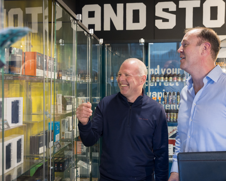 Two men, in business attire, chat in a shop while looking at glass display cabinets.
