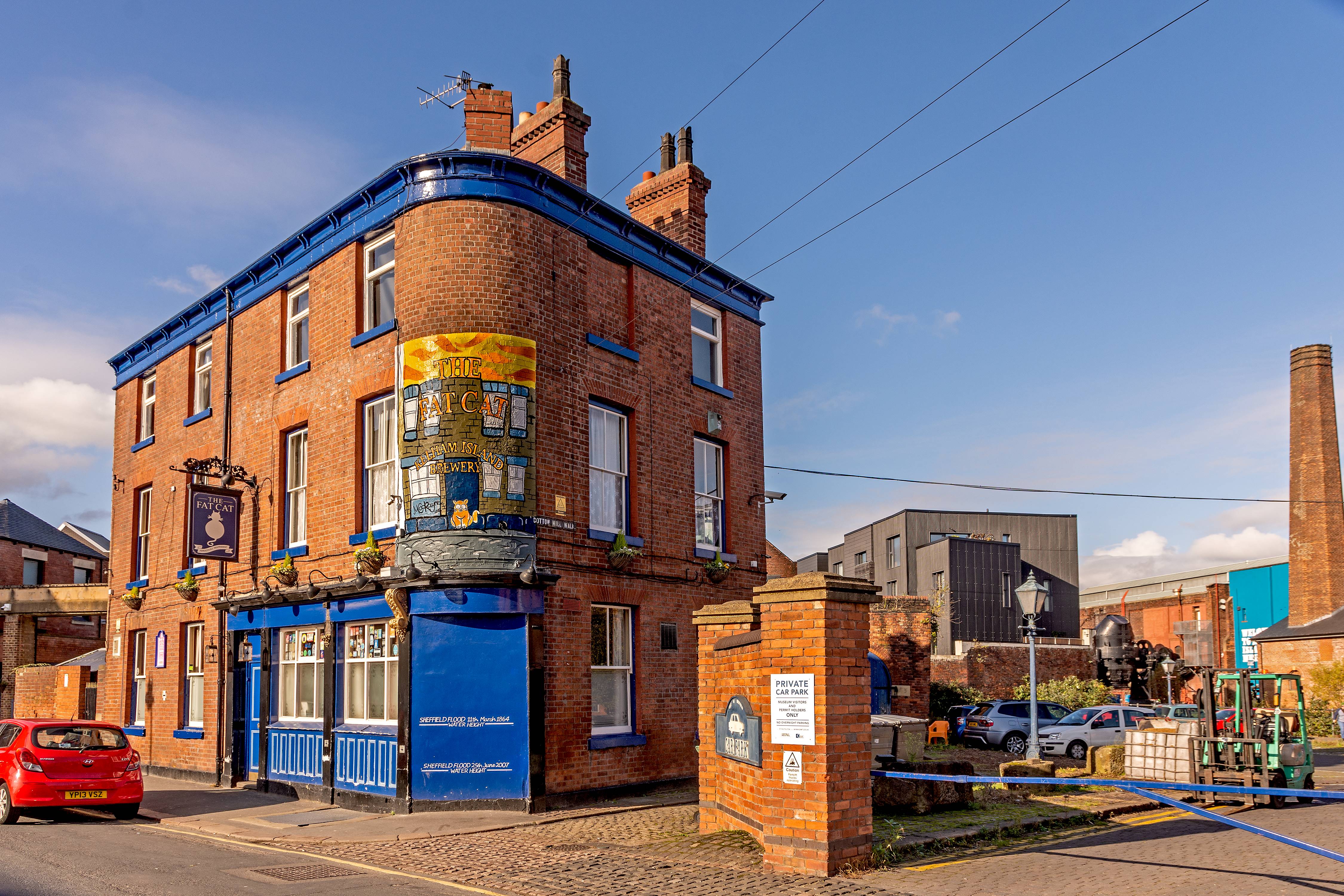 The Fat Cat pub in Kelham Island - Historic red-brick pub building with bright blue trim on a street corner, featuring a large painted sign reading ‘The Victoria Junction Inn.’ A red car is parked nearby, and industrial-style buildings and a tall chimney are visible in the background under a clear blue sky.