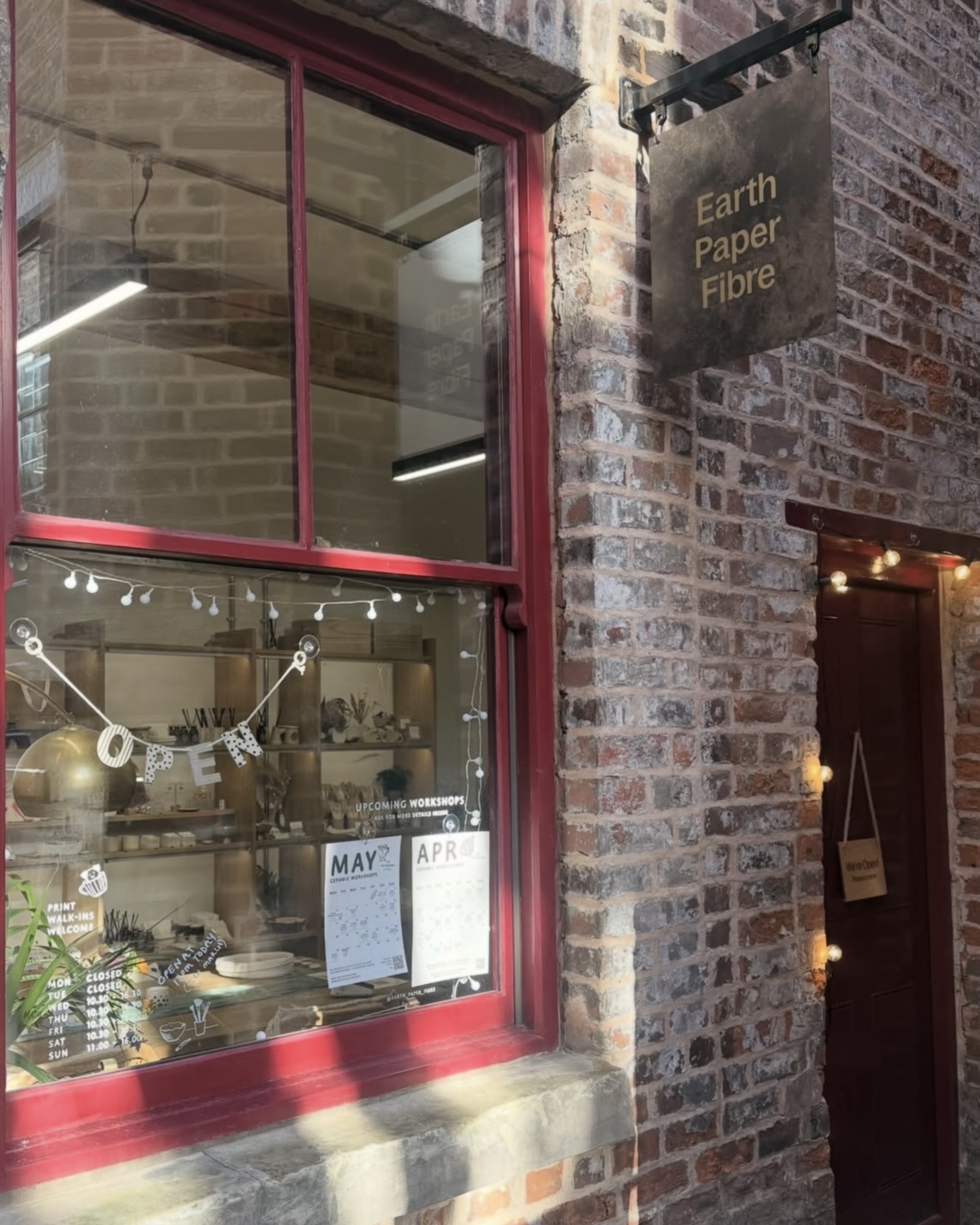 Exterior of a brick‑fronted shop with a red‑framed window displaying ceramics, and a hanging sign reading “Earth Paper Fibre” above the entrance.