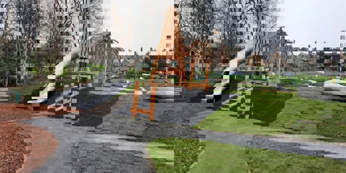 Playground with a large wooden-framed structure and a long, curved metal slide. The area is surrounded by grass, pathways, and leafless trees. Stone buildings and fencing are visible in the background under an overcast sky.