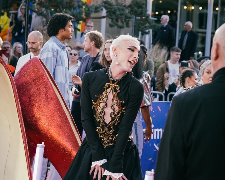 A person in an elaborate black outfit with gold detailing poses on a red carpet. In the background, a crowd watches and takes photos under festive string lights. Large decorative high-heeled shoe props are visible in the foreground.