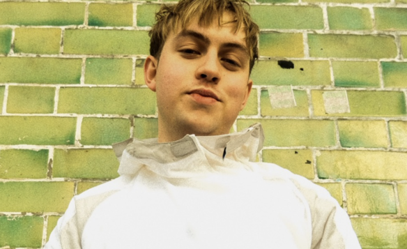 Person wearing a light-coloured jacket standing against a wall made of green bricks with varying shades and weathered texture.